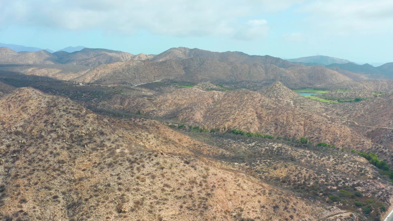 Third frame holds on the Baja California Sur mountain landscape, with tan ridges across the foreground, a small bright green valley area at right, hazy mountains behind, and no visible people.