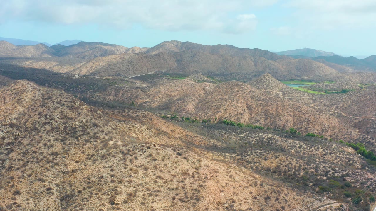 Fourth frame shows an elevated wide view near Marquis Los Cabos, with the foreground ridge lower in frame, multiple dry hills across the midground, a green valley patch to the right, and no visible people.