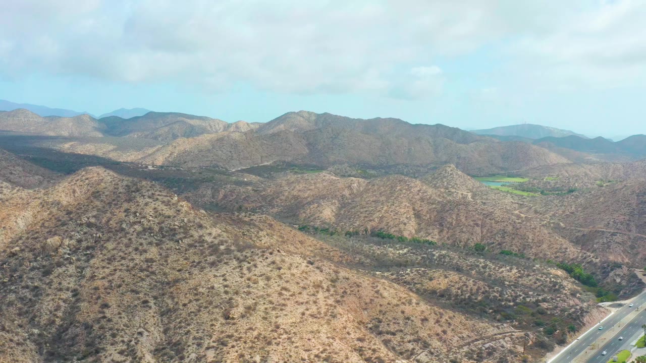 Opening frame over Marquis Los Cabos, Baja California Sur, with dry rounded hills in front, distant ridges across the horizon, a highway at lower right, green valley patches, and no visible people.
