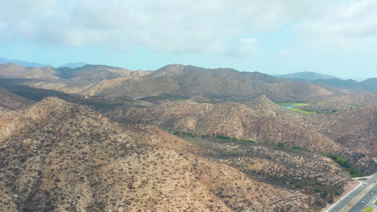 Wide aerial view near Marquis Los Cabos with a rounded hill in the foreground, rocky brown terrain across the middle distance, a road entering at lower right, distant ridgelines, and no visible people.