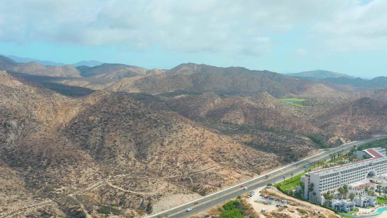 Aerial frame over Marquis Los Cabos, Baja California Sur, with dry hills around a paved highway, small cars, a white hotel building, green grounds, and no visible people.