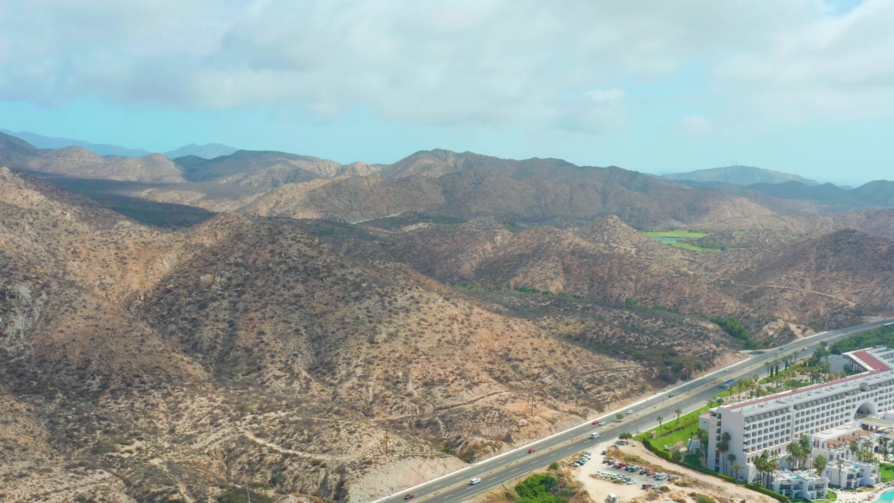 Aerial view of Marquis Los Cabos with layered tan hills, a road cutting through the lower right, small cars, the edge of a white hotel, pool structures, and no visible people.