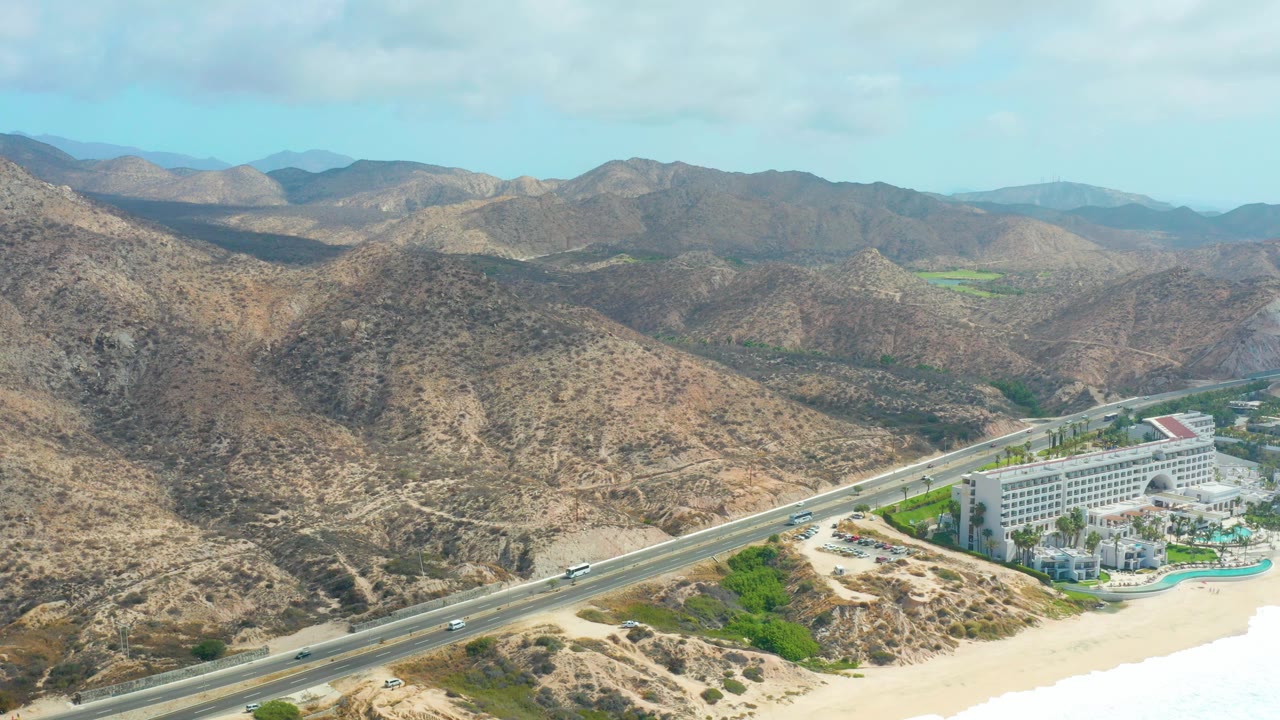 Opening aerial frame over Marquis Los Cabos with a coastal highway, a white resort beside the sandy beach, arid mountain ridges in the background, and no visible people.