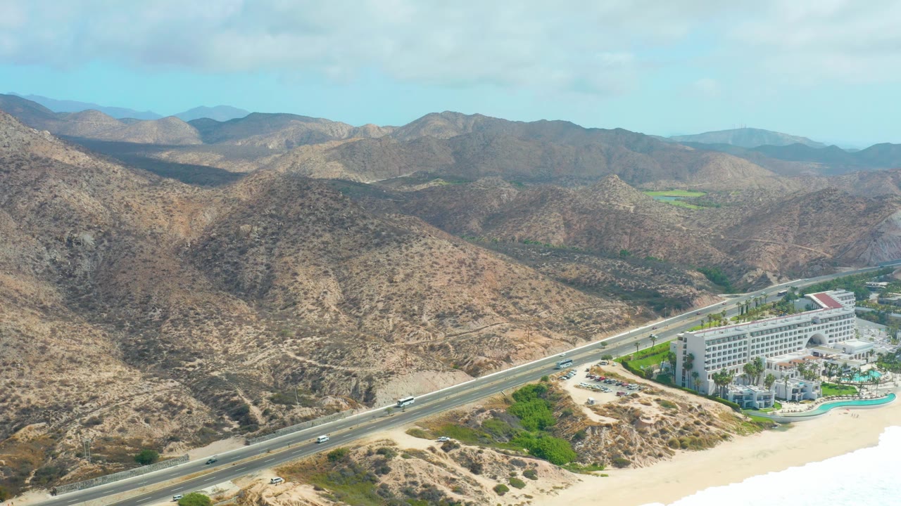 Aerial frame over Marquis Los Cabos showing small vehicles on the coastal highway, the resort building near the lower right, sandy shoreline, dry hills, and no visible people.