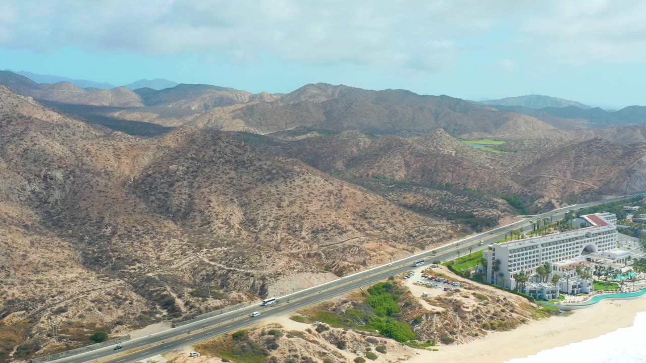 Wide aerial frame in Marquis Los Cabos with the resort lower right, the highway running diagonally across the coast, layered brown mountains behind, and no visible people.