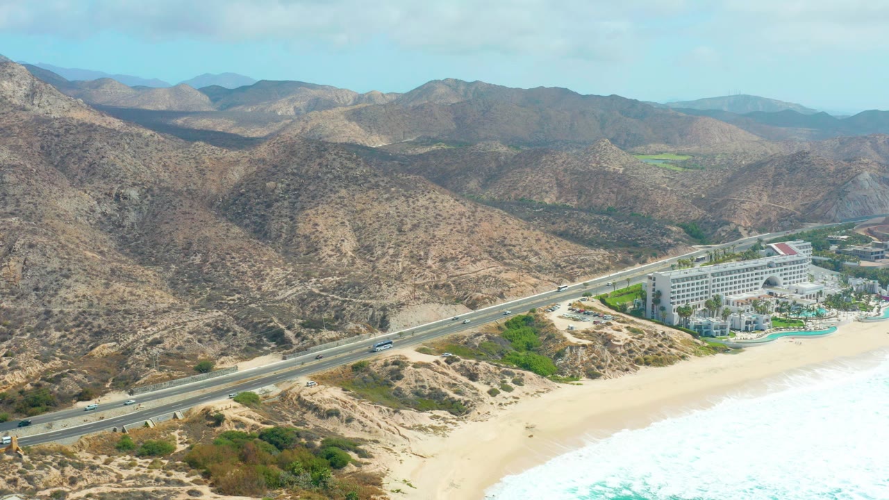 Aerial view at Marquis Los Cabos, Baja California Sur, with a coastal highway crossing the lower frame, a resort complex beside pale sand and surf, dry mountains behind it, and no visible people.