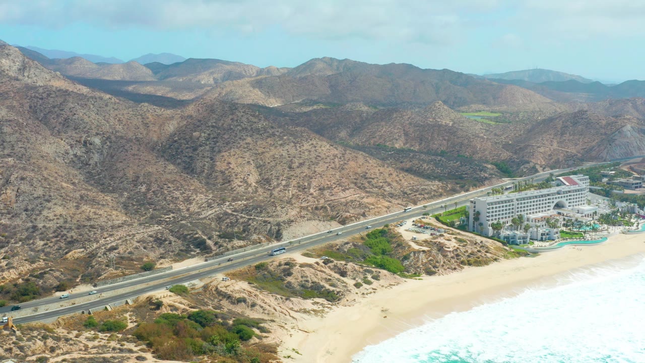 A one-second view of Marquis Los Cabos shows vehicles on the coastal road, white surf washing the beach, a large resort building near the shore, brown mountain slopes, and no visible people.