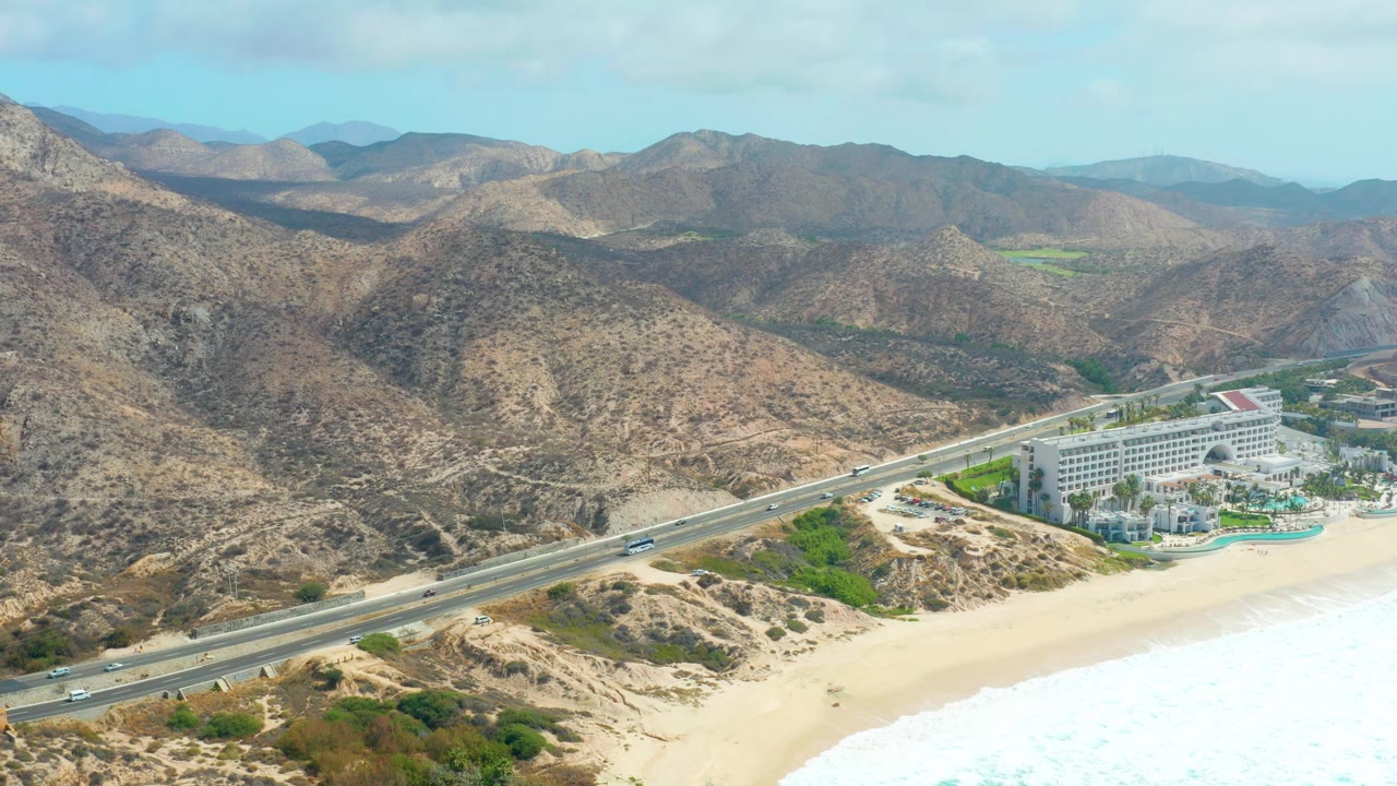 The aerial frame at Marquis Los Cabos shows the highway cutting between dry coastal hills and the beach, with vehicles visible, surf at the lower right, resort structures near the shore, and no visible people.