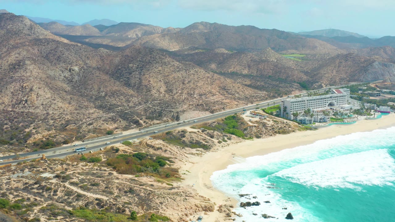 Aerial view at 0:01 over Marquis Los Cabos, with the highway running across scrubby dunes, the resort closer to center, surf washing onto the beach, and no visible people.