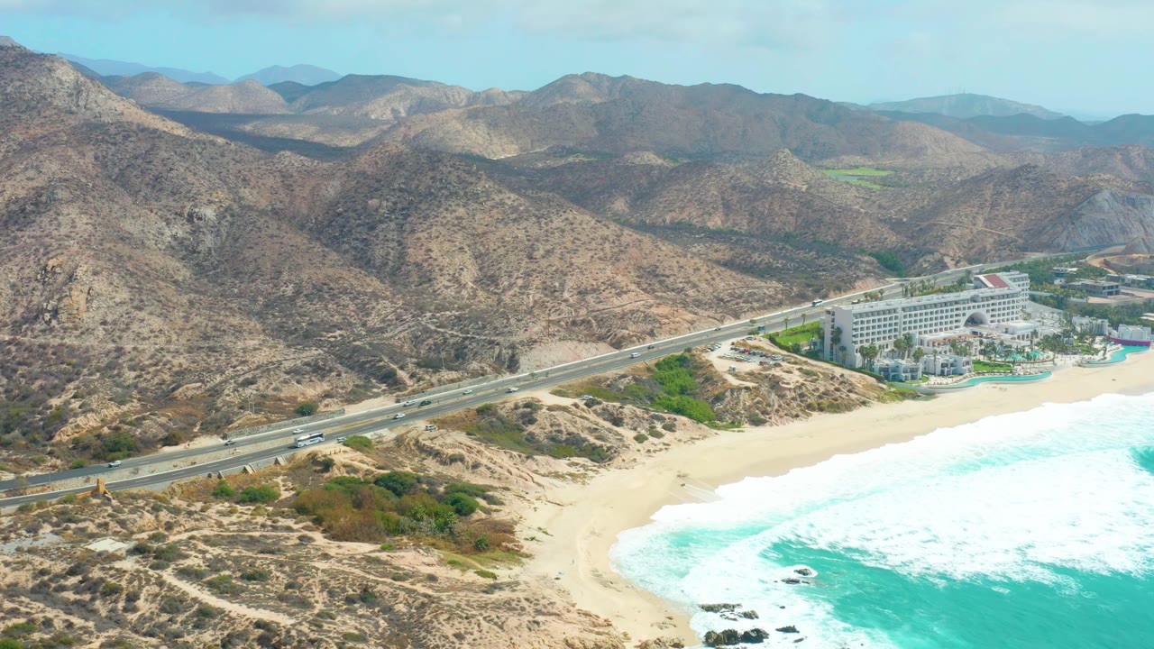Mid-clip aerial frame over Marquis Los Cabos, with the highway spanning the lower hillside, surf pushing onto the sand, the resort at the coast, and no visible people.