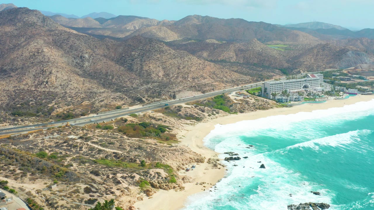 At 0:00, an aerial view over Marquis Los Cabos shows a coastal highway beside pale sand and turquoise waves, with dry rocky mountains behind and no visible people.