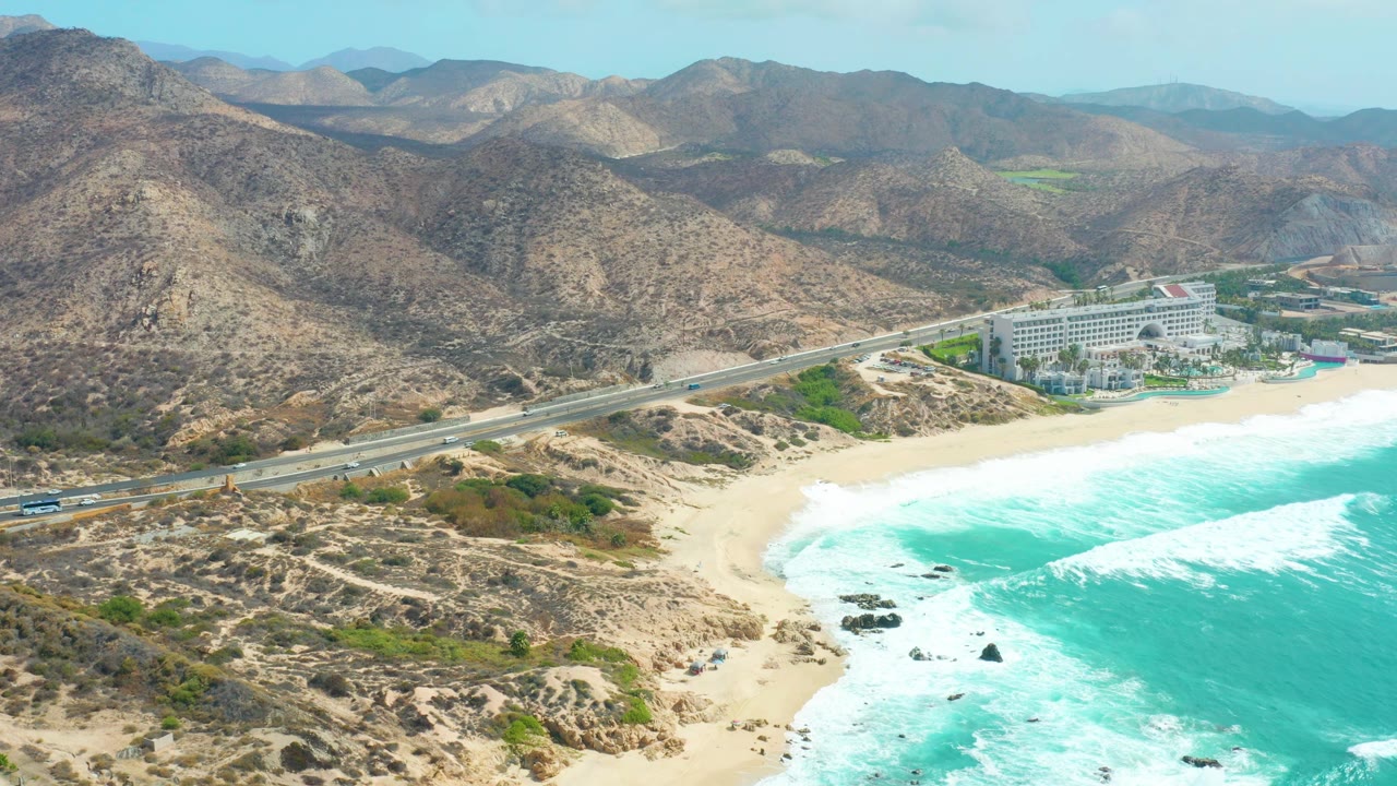 Aerial view of highway traffic beside beach surf and rocky desert hills.
