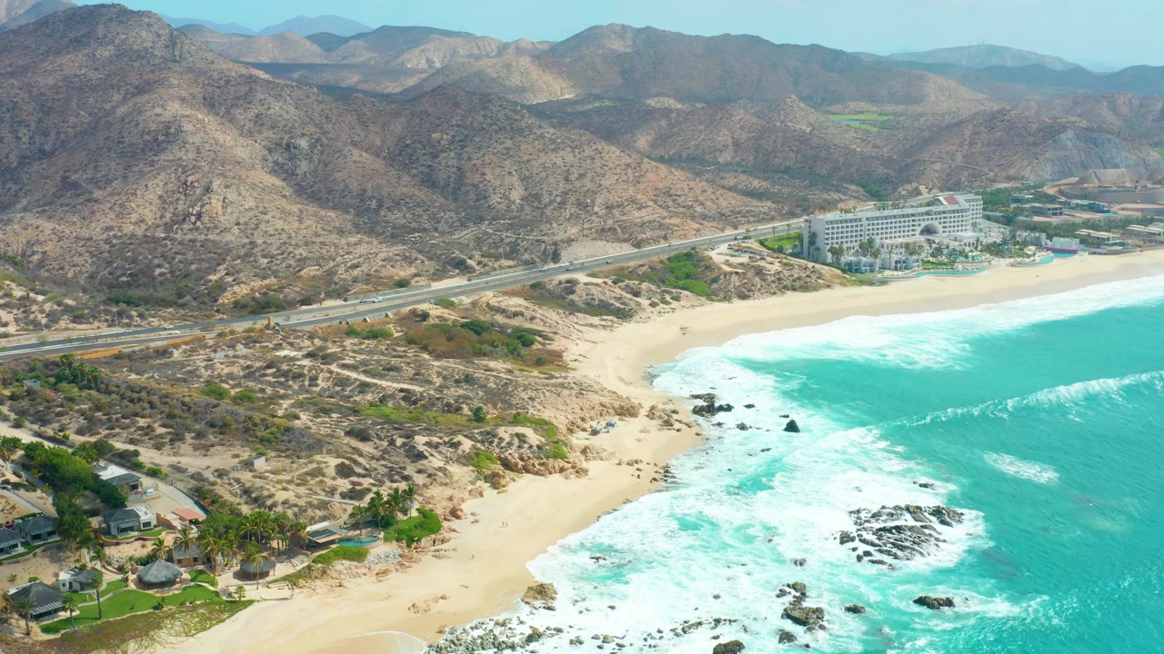 Aerial frame over Marquis Los Cabos showing turquoise surf breaking on sand, a coastal road crossing the midground, arid hills behind it, a large beachfront building, and no visible people.