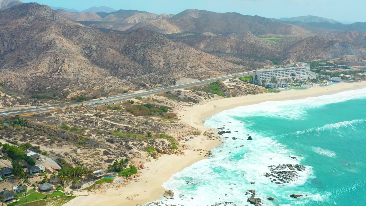 High aerial frame of Marquis Los Cabos with the surf line curving around a sandy cove, a road and large beachfront building beyond it, arid mountains, and no visible people.