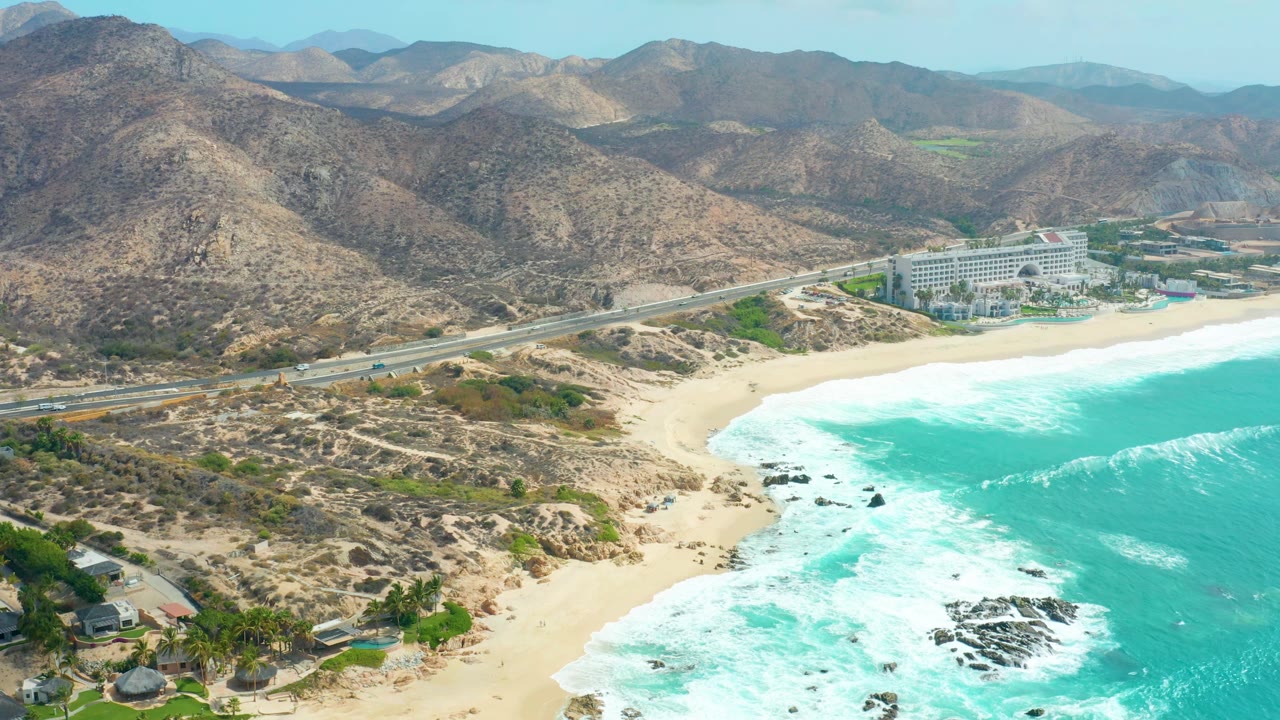 Aerial beach scene with surf, road, vehicles, hillside, and coastal structures.