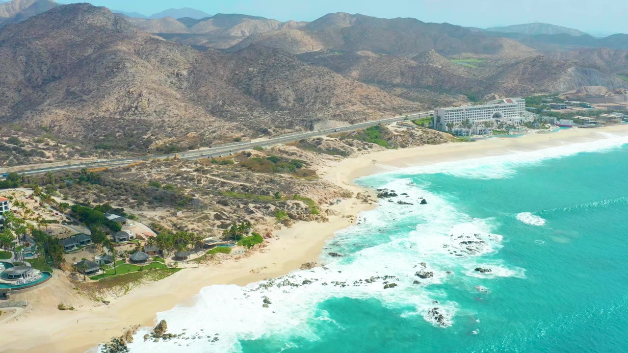 Opening aerial frame over Marquis Los Cabos showing turquoise waves, white foam, sandy beach, dark rocks, coastal buildings, a highway, dry mountains, and no visible people.