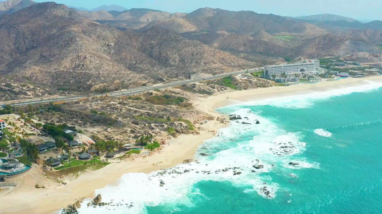 Aerial frame at Marquis Los Cabos with a broader band of white surf spreading across the rocks, sandy beach below, highway and buildings behind it, mountains in the distance, and no visible people.