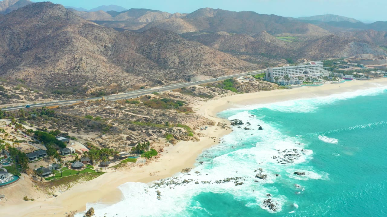 Aerial view of foamy waves against rocks near a sandy beach and highway.