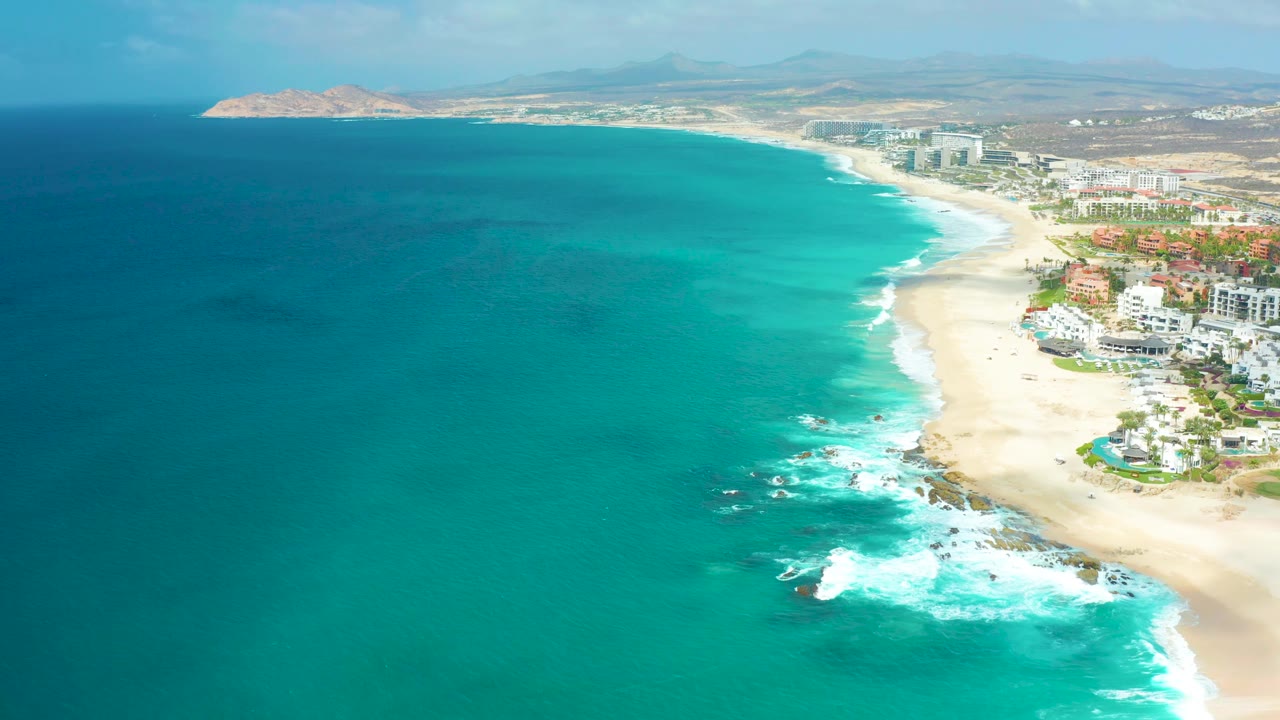 Marquis Los Cabos coastline seen from above, with white waves reaching the rocky shore, pale sand curving into the distance, buildings on the right, and no visible people.
