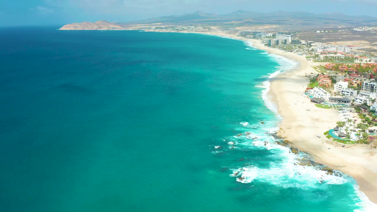 High aerial view over Marquis Los Cabos with darker water at left, bright turquoise shallows near shore, white surf below, beach and buildings to the right, and no visible people.