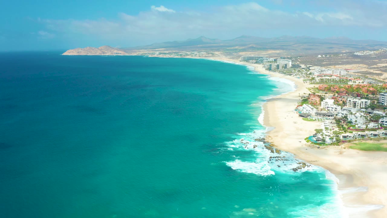 Opening frame over Marquis Los Cabos shows deep blue water shifting to turquoise near a sandy beach, with white surf, coastal buildings on the right, distant land, and no visible people.