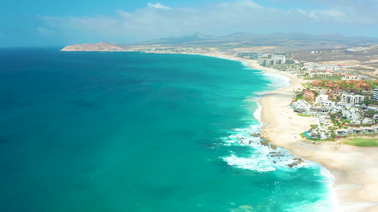 At 0:02, turquoise water dominates the frame while a band of white foam breaks near the sandy beach, with Marquis Los Cabos coastal buildings partly visible on the right and no visible people.