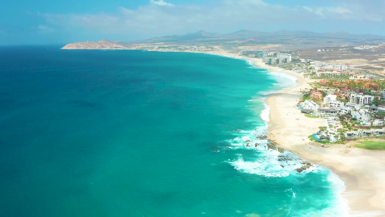 Aerial beach coast with turquoise sea, white waves, buildings, and distant land.