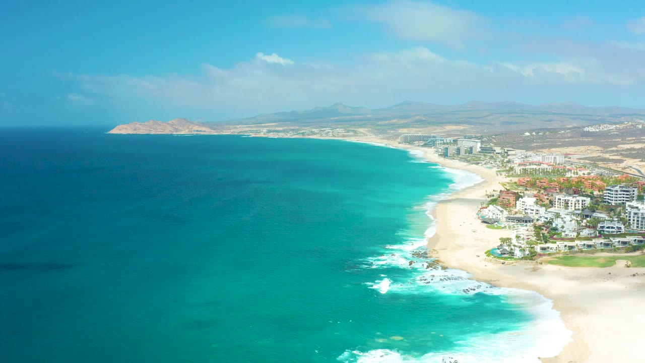 Aerial view at Marquis Los Cabos with turquoise ocean on the left, a curving sandy beach on the right, coastal buildings near the shoreline, distant inland terrain, and no visible people.