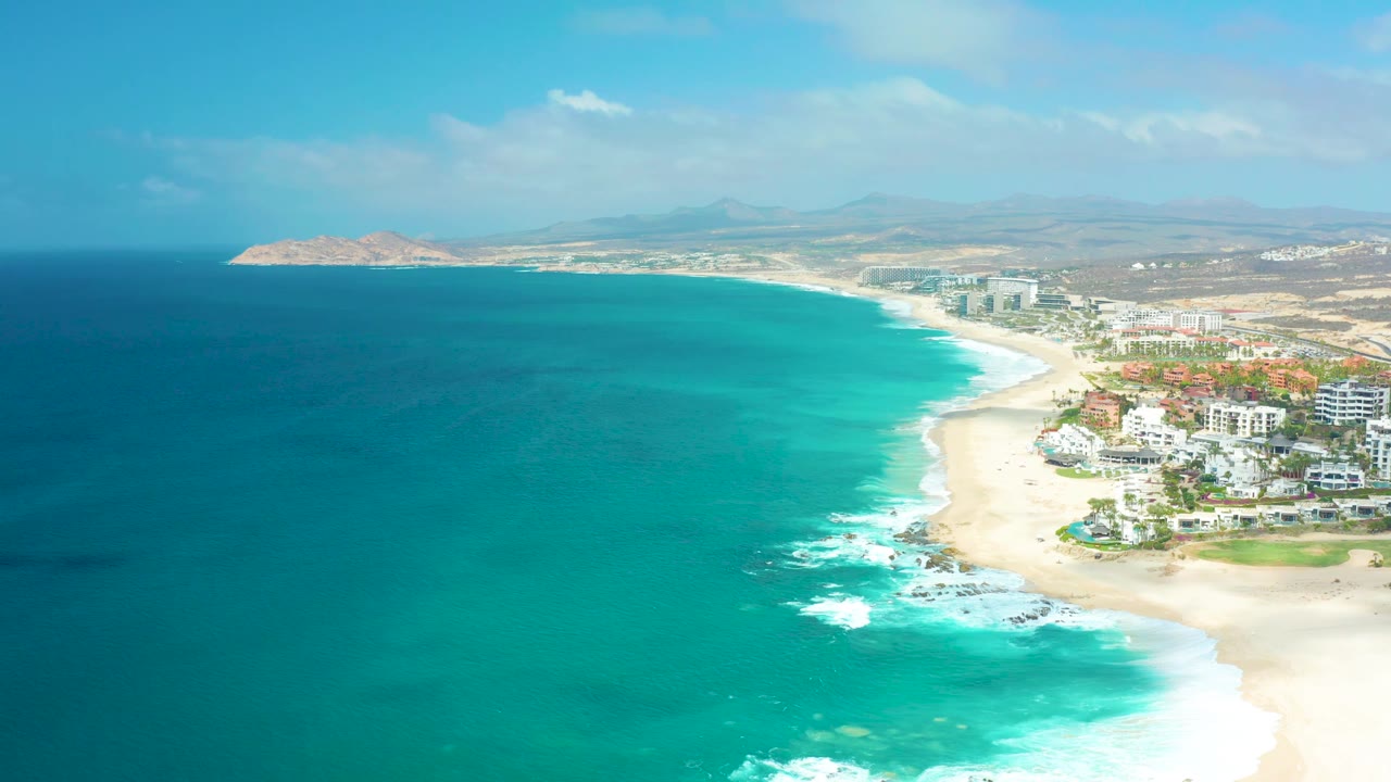 Ocean and sandy shoreline with white waves near coastal buildings.