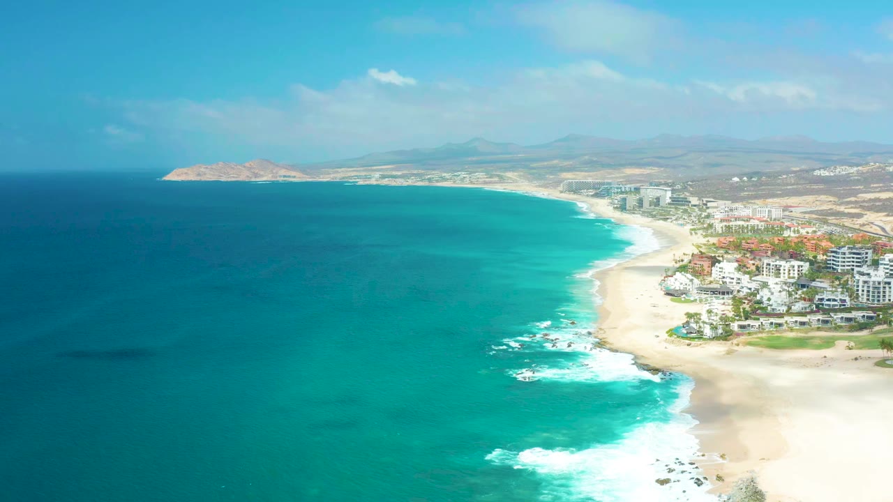 Opening aerial frame over Marquis Los Cabos showing turquoise ocean on the left, white surf on the beach, beachfront buildings on the right, hazy inland hills, and no visible people.
