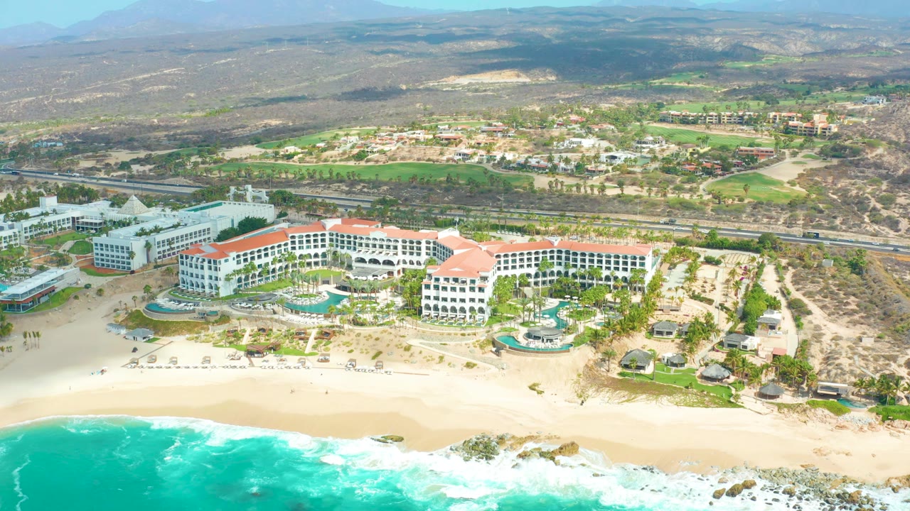 Wide aerial frame over Marquis Los Cabos, with turquoise ocean and sandy beach in front of white resort buildings, pools, palms, dry hills, and distant mountains, with no visible people.