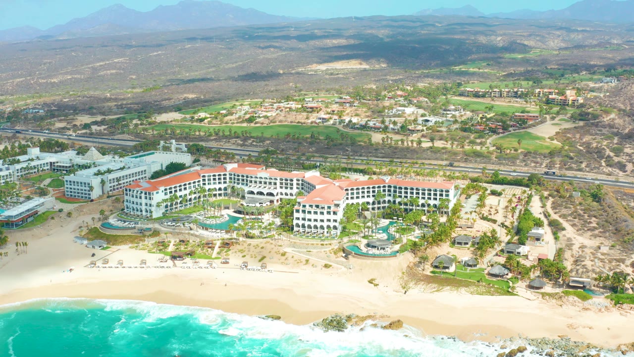Marquis Los Cabos appears from a high coastal angle, with curved white hotel buildings, circular pool areas, palms, a sandy beach, turquoise wave wash, inland green grounds, desert hills, and no visible people.