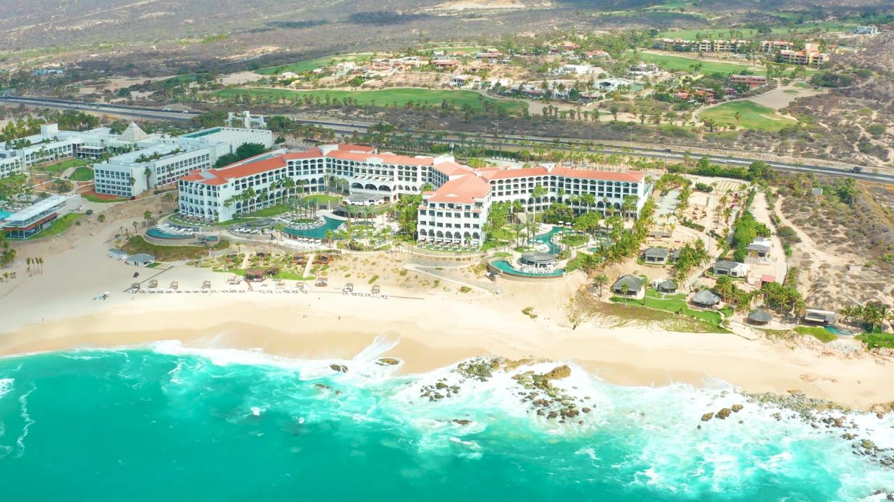 Aerial view over Marquis Los Cabos as a white wave line spreads across the turquoise water below the sandy beach and resort complex, with no visible people.
