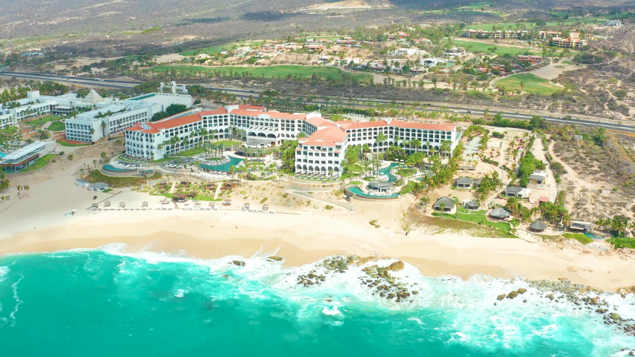 Aerial frame of Marquis Los Cabos with resort pools and white buildings centered above the beach while a broad turquoise band of ocean fills the foreground, with no visible people.