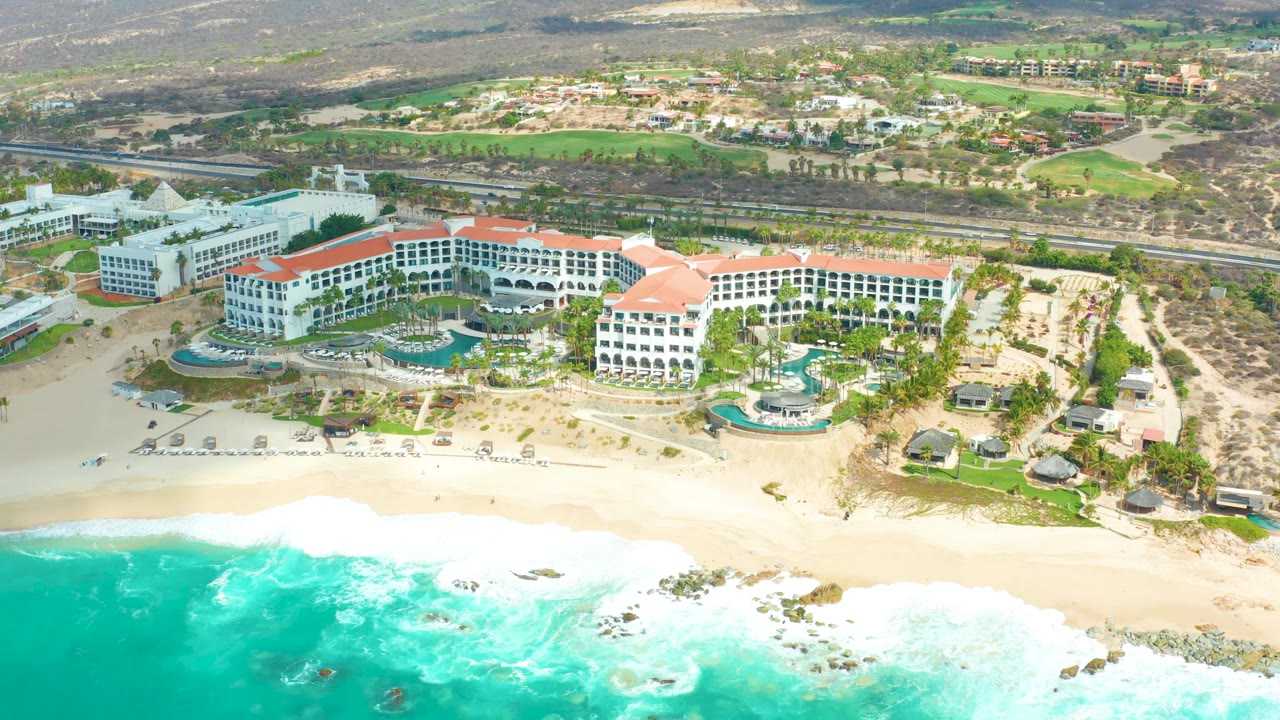 Aerial frame at Marquis Los Cabos showing turquoise surf breaking across the sandy beach below white resort buildings with red roofs, pools, palms, and no visible people.