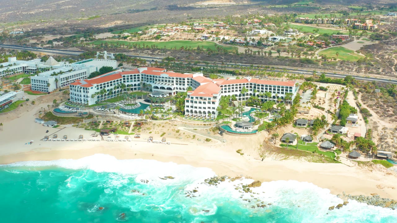 Aerial view of the Marquis Los Cabos beachfront as white foam spreads closer to the sand, with the resort complex, pools, road, and hillside remaining in the background and no visible people.