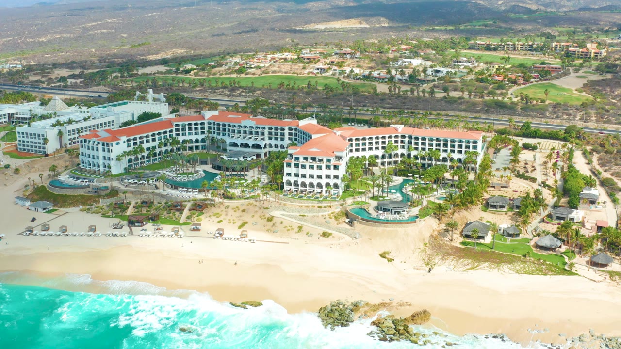 At 0:00, an aerial view shows Marquis Los Cabos with turquoise surf in front, a sandy beach, white red-roofed resort buildings, pools, a road, inland greenery, hills, and tiny beachgoers visible.
