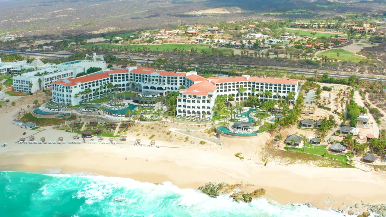 At 0:02, the frame shows Marquis Los Cabos from above with more bright surf visible at the lower left, red-tiled roof sections across the hotel, pools near the beach, and inland roads and hills beyond.