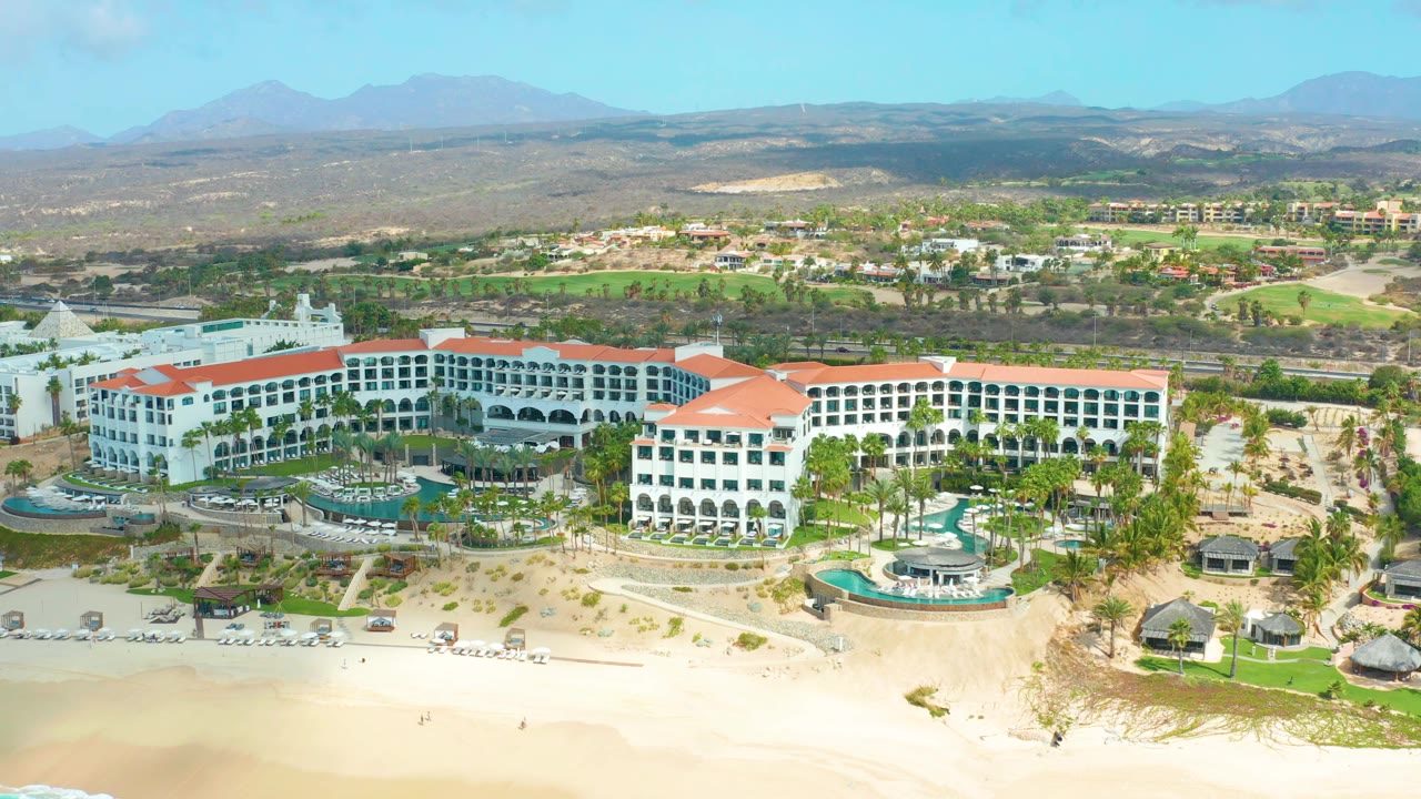Wide aerial frame of Marquis Los Cabos showing white resort buildings with red roofs above a sandy beach, landscaped pools, inland greenery, dry hills, distant mountains, and no visible people.