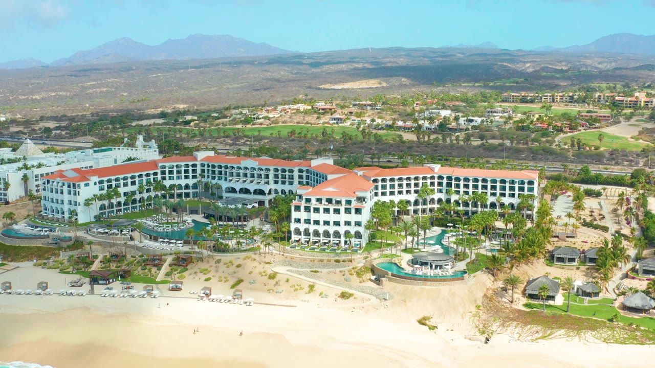 Aerial resort view with beach frontage, pools, palm landscaping, and inland mountains.