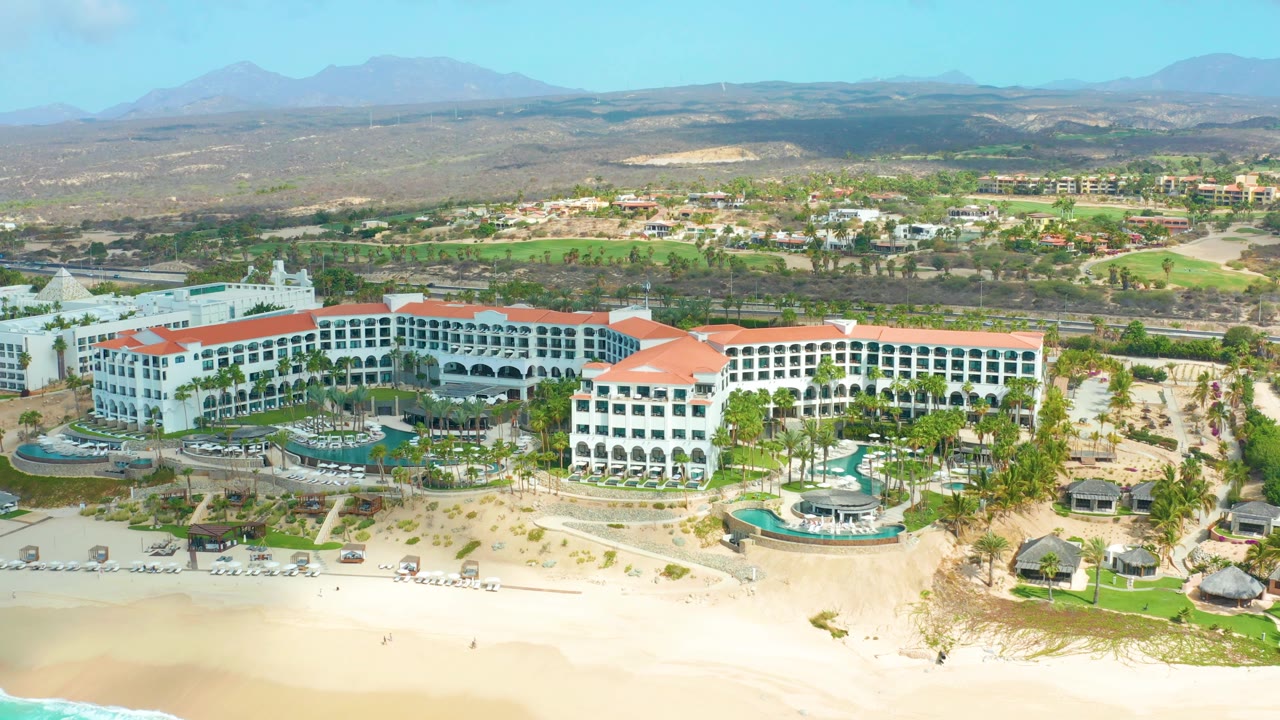 Wide aerial frame of Marquis Los Cabos with white arched resort facades, red roofs, pool terraces, beach umbrellas or loungers near the sand, inland greenery, mountains, and no visible people.