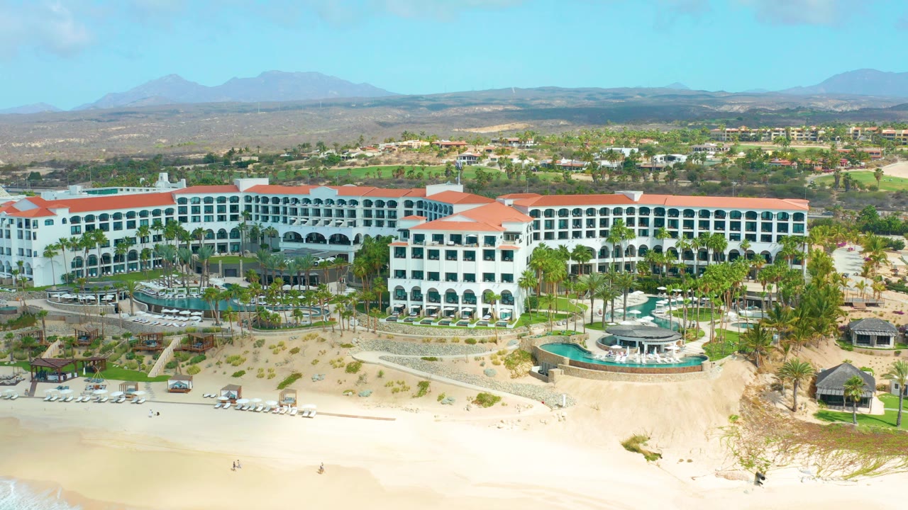 Opening aerial frame of Marquis Los Cabos showing the white resort block, red roofs, sandy beach foreground, palm landscaping, distant mountains, and no recognizable people visible.