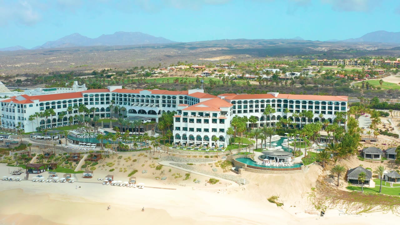 Aerial pan over the resort with more inland landscape and golf-like green areas visible behind it.