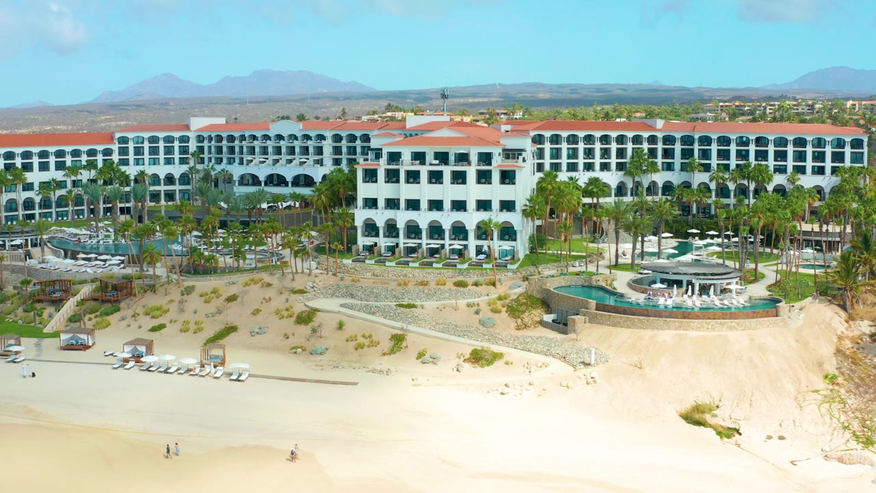 Opening aerial frame of Marquis Los Cabos with a white resort, red tile roofs, palm-lined grounds, pale sand, distant mountains, and tiny distant beachgoers.