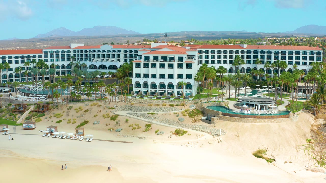 Early aerial frame of Marquis Los Cabos showing the central white hotel block, curved pool terrace, palms, beach loungers, pale sand, and mountains beyond.