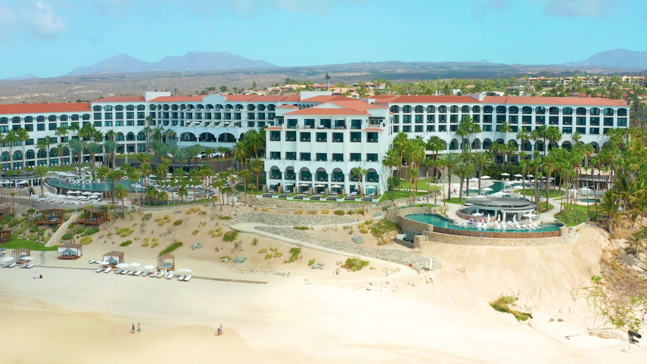 Aerial frame of Marquis Los Cabos with long white hotel wings, repeated balcony arches, landscaped palms, a bright sandy beach, and a blue mountain horizon.