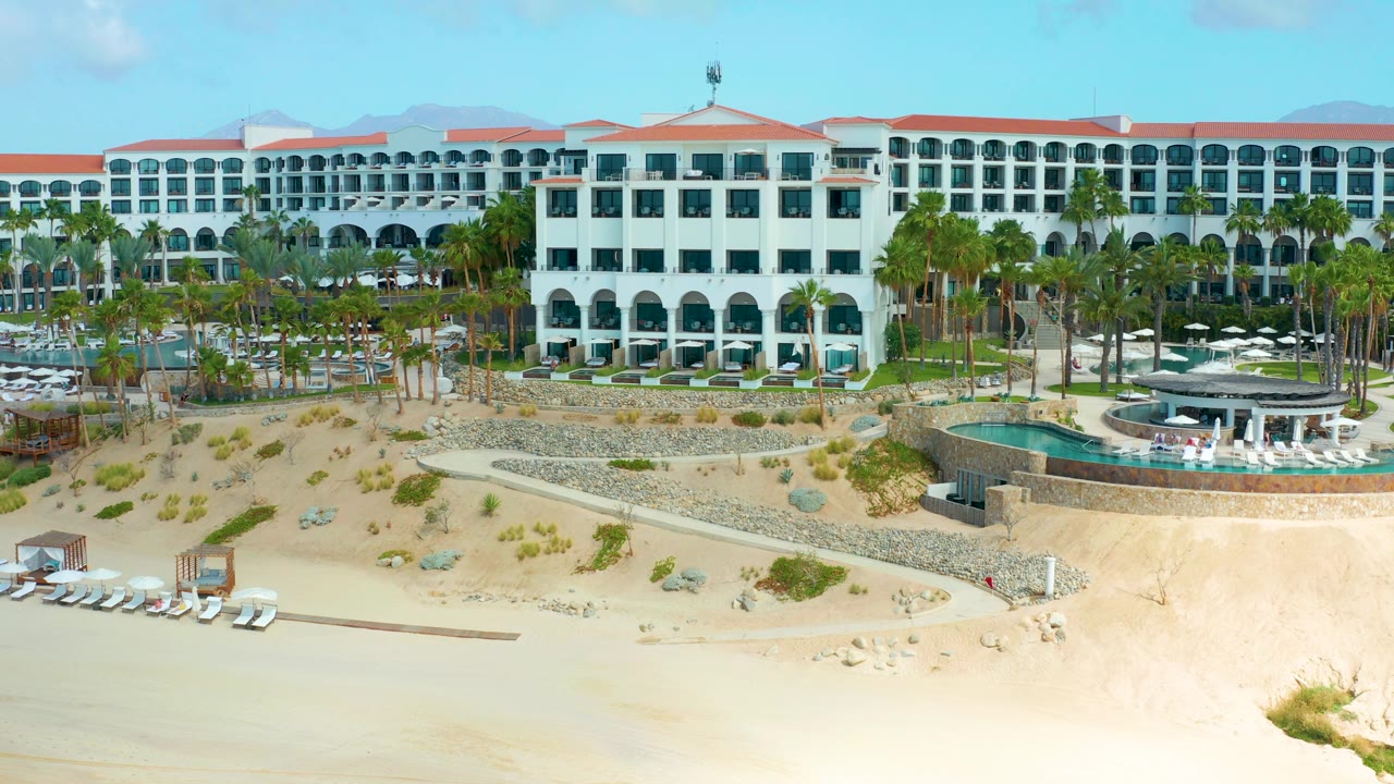 Elevated view of beachfront hotel, palms, pool terraces, dunes, and beach chairs.