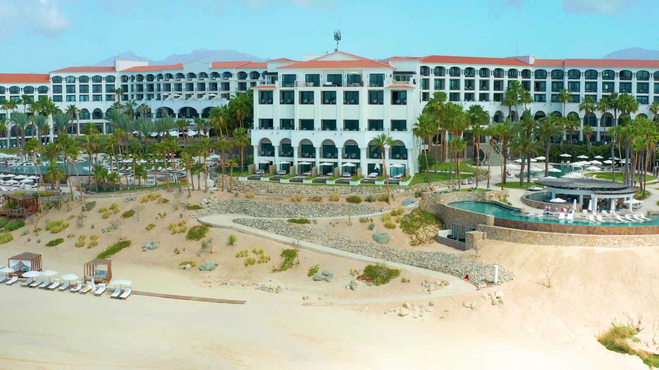 A similar elevated view shows the Marquis Los Cabos hotel stretched across the background, with the central white block, palms, pool deck, dunes, and beach below.