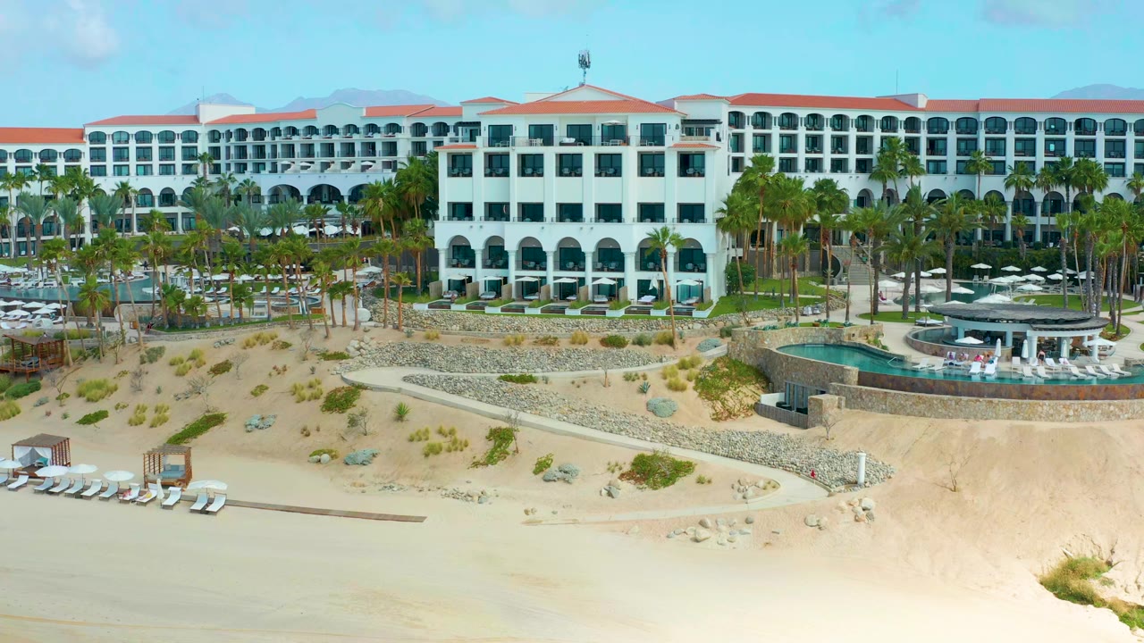 White resort buildings and palm-lined pool area above a sandy beach with lounge chairs.