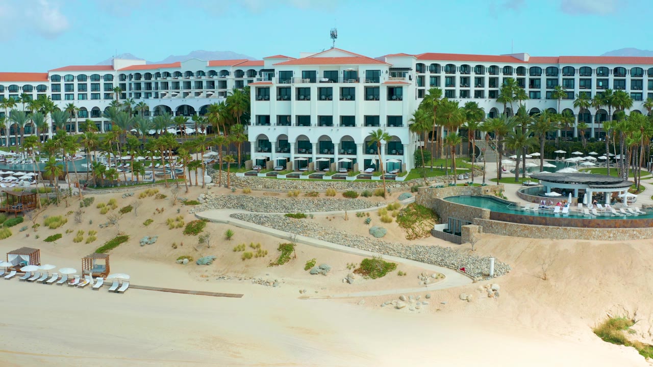 At 0:02, the Marquis Los Cabos exterior shows repeating arched windows, red rooflines, palms around the pool area, beach lounge chairs near the sand, and no visible people.
